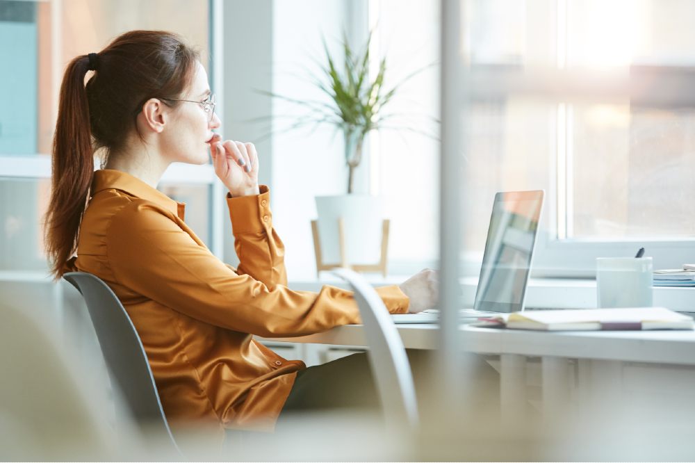 Red-haired woman in a yellow shirt working on her laptop while thinking and looking out of the window