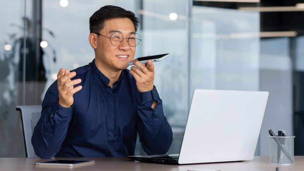 Man wearing a blue shirt talking into his phone via speaker in front of his laptop